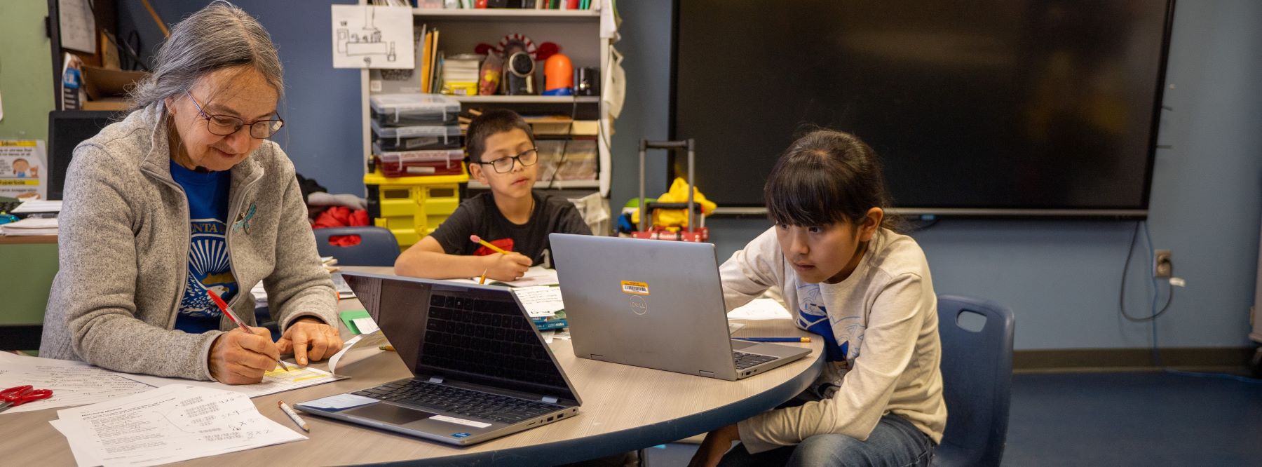Teacher and two students sitting at a table using their laptops and focused on their screens.